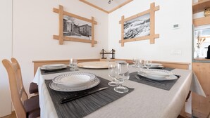 A dining area with a table set for four, featuring white plates with blue patterns, clear wine glasses, and a knife. The room has wooden beams on the walls and two framed pictures.