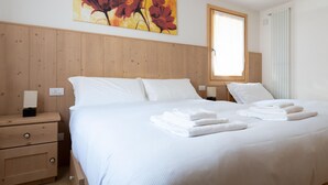 A neatly made bed with white linens and folded towels, a wooden nightstand with a lamp, and a window with blinds.