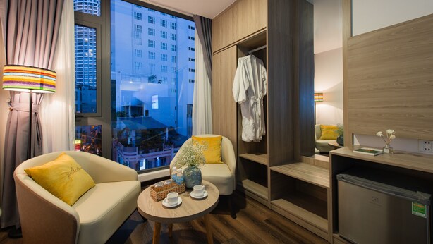 Ein Hotelzimmer mit einer Couch, einem kleinen Tisch mit Tasse und Wasserflaschen, einer Holzkabine und Blick auf eine Stadtlandschaft durch große Fenster.