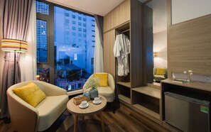 A hotel room with a sofa, a small table with a cup and water bottles, a wooden cabinet, and a view of a cityscape through large windows. - Holi Anna Boutique (Nha Trang)