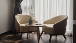 Ein Zimmer mit zwei beigen Sesseln, einem kleinen runden Tisch mit Vase und Buch, einer hohen Stehlampe mit gestreifter Lampenschirm und durchsichtigen Vorhängen.