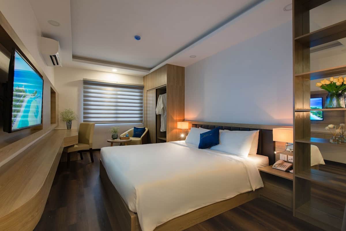Une chambre d’hôtel avec un grand lit, une télévision, un bureau avec une lampe et une étagère ornée de fleurs.