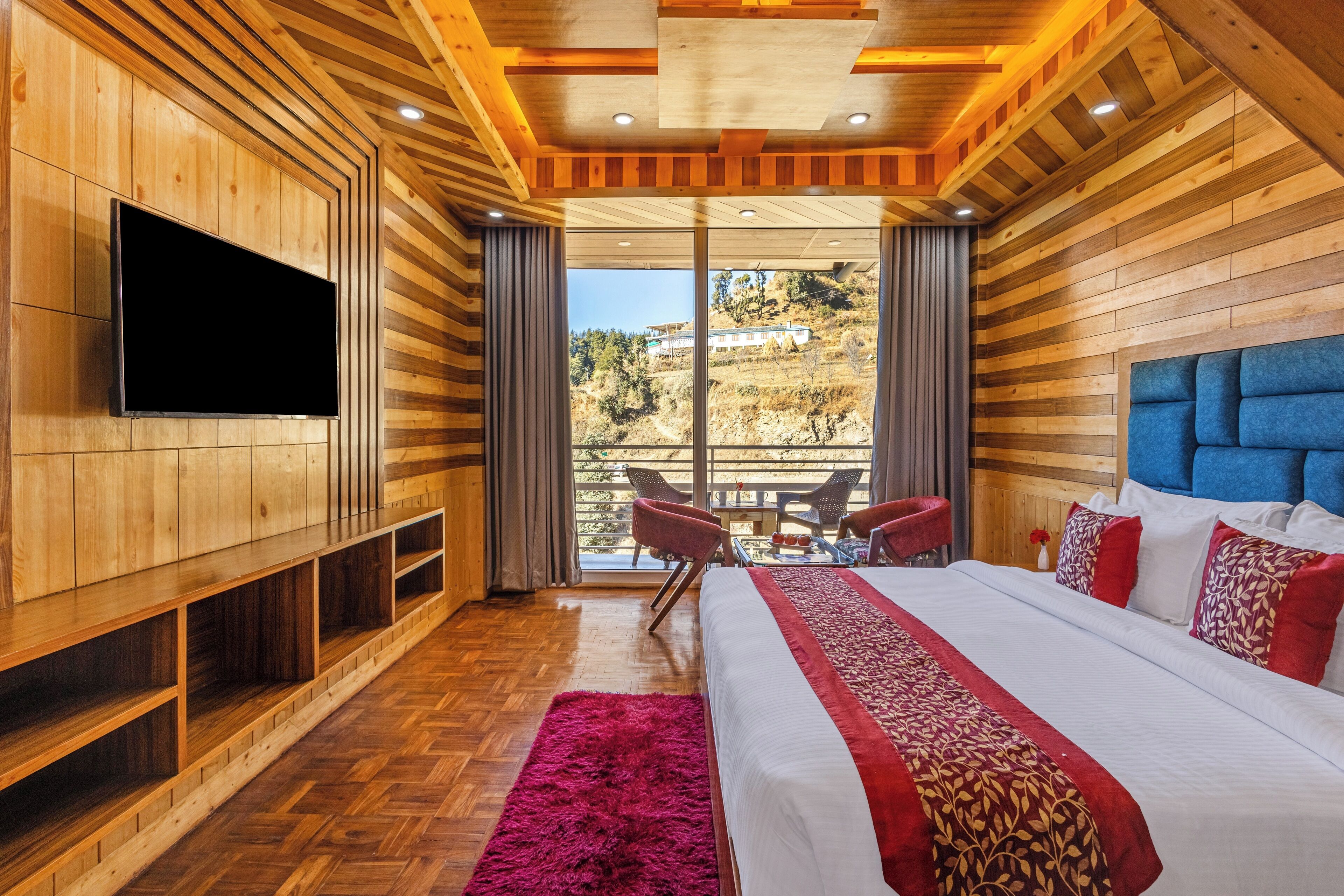 A modern bedroom with a large bed, a flat-screen TV, wooden paneling, and a balcony with outdoor seating.