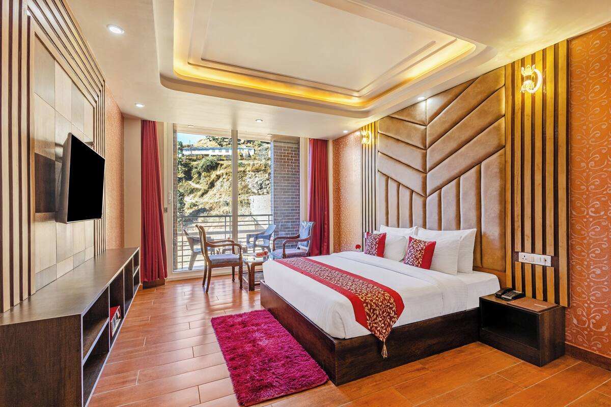 A modern hotel room with a large bed, a flat-screen TV, a balcony with chairs, and a view of the outdoors.