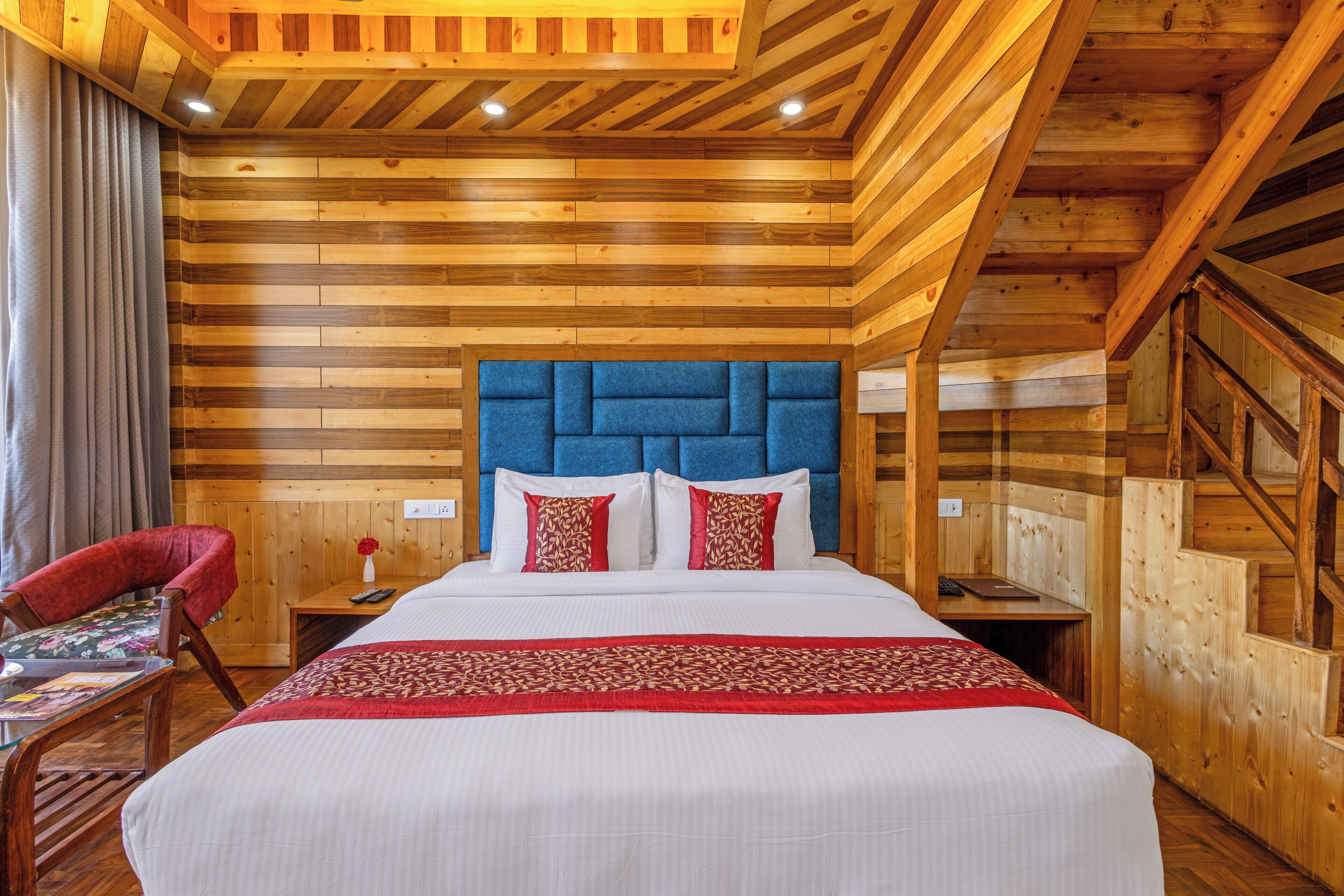 Una habitación con paneles de madera, una cama grande, una silla roja y una mesita con un jarrón de flores.