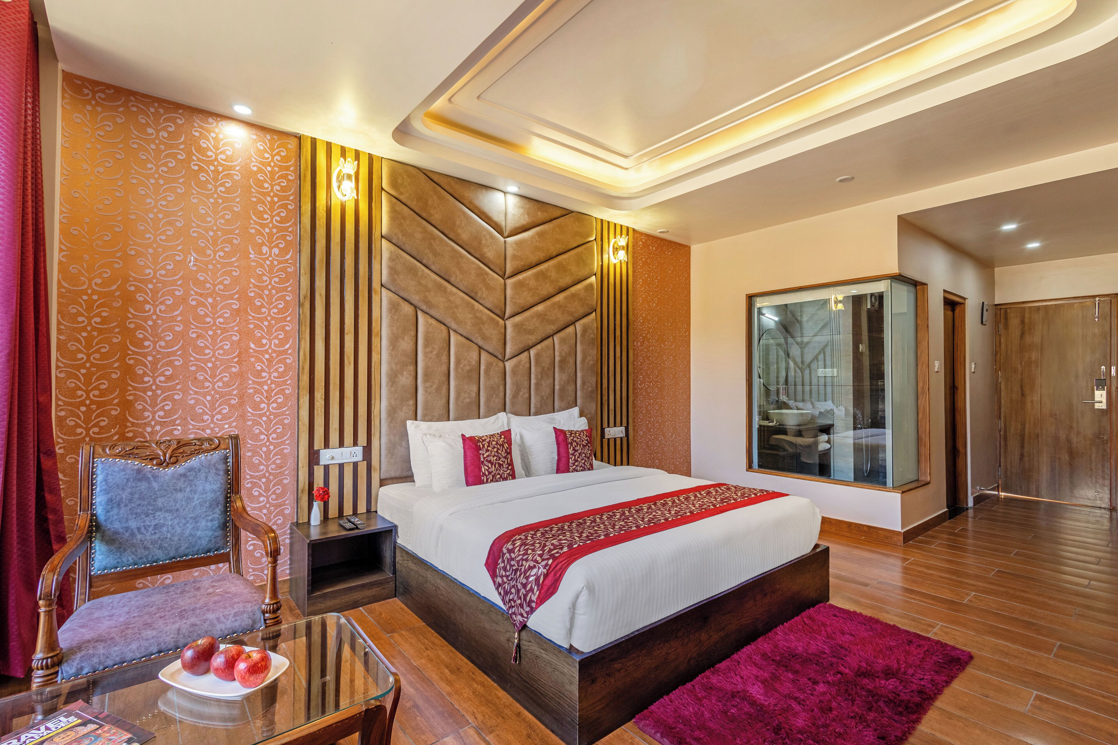 A modern hotel room with a large bed, a glass shower, a wooden wardrobe, and a plush armchair.