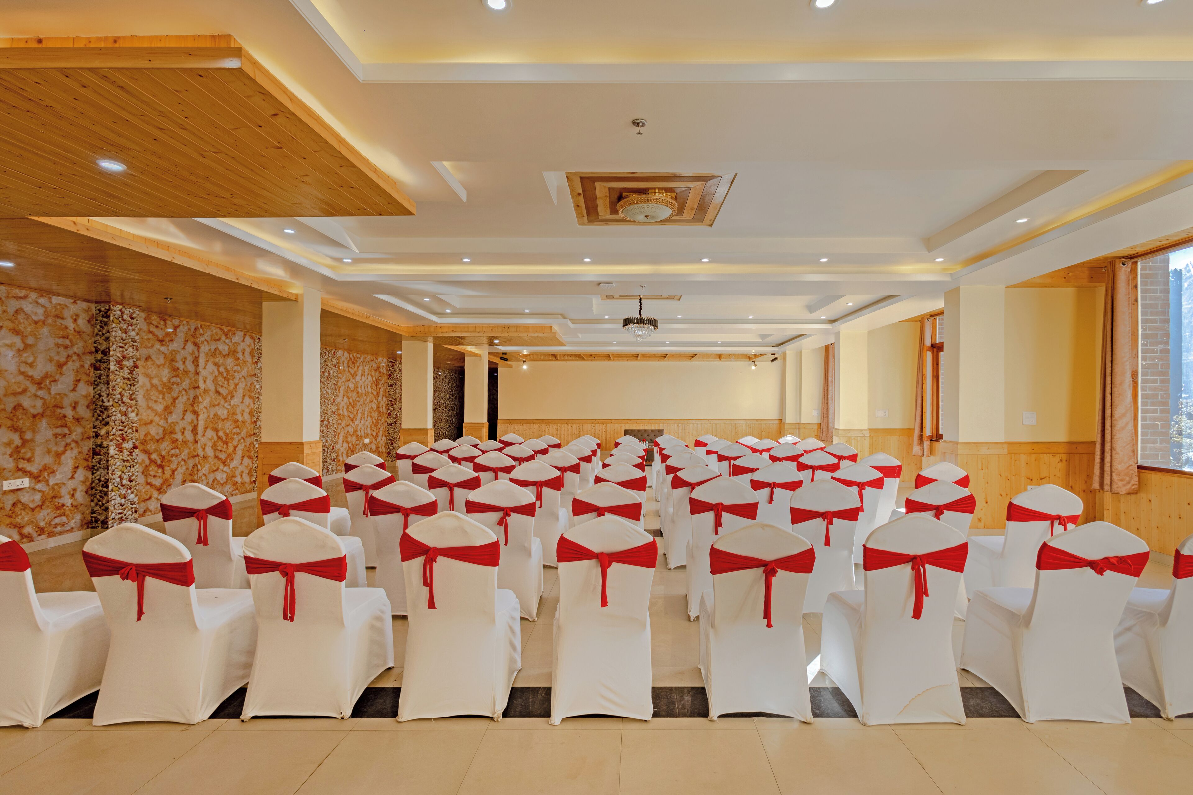 Un salón de conferencias con hileras de sillas blancas con cintas rojas, paneles de madera en las paredes e iluminación empotrada en el techo.