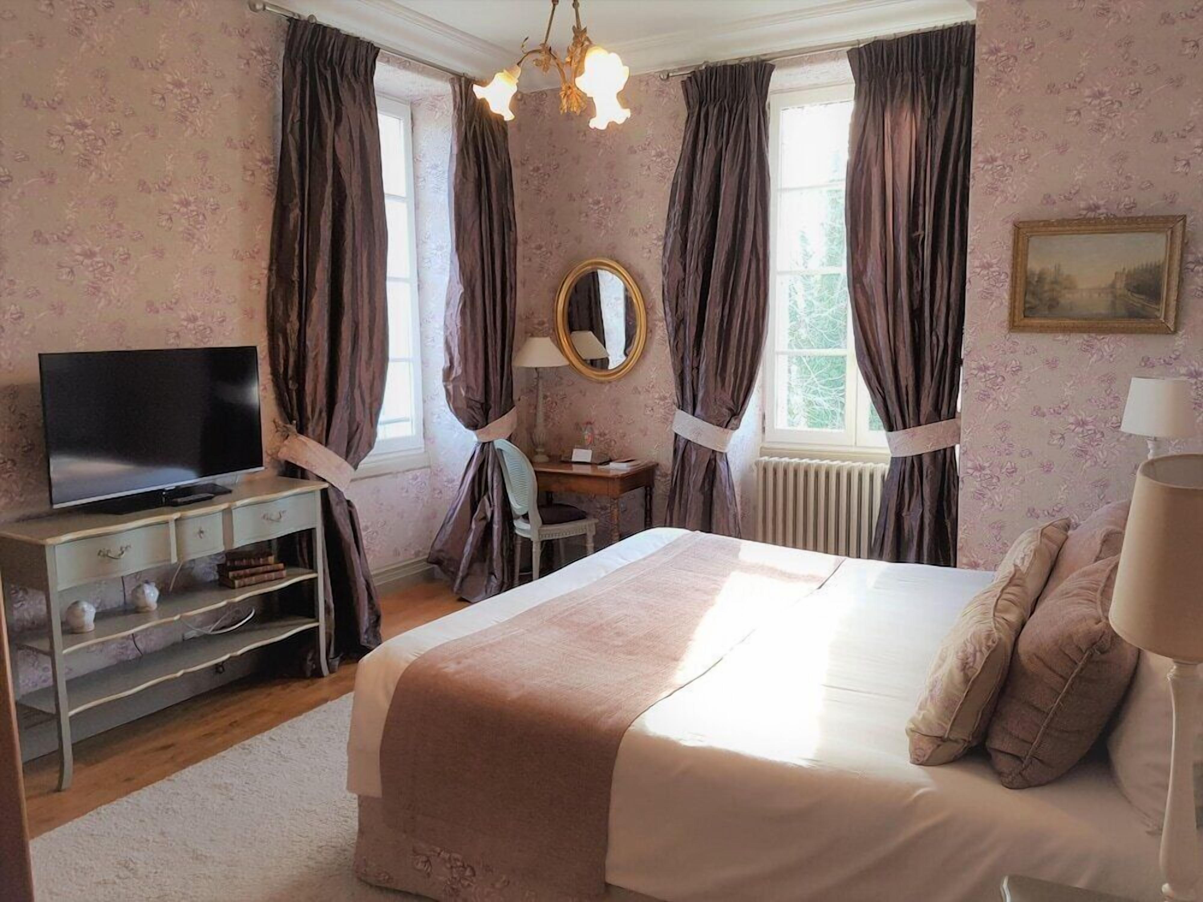 Een slaapkamer met een bed, een televisie op een dressoir, een spiegel en een raam met gordijnen.