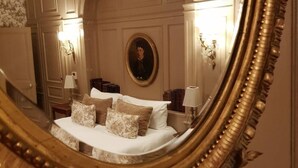 Een slaapkamer met een grote, versierde spiegel die een bed met witte lakens en veel kussens weerspiegelt.