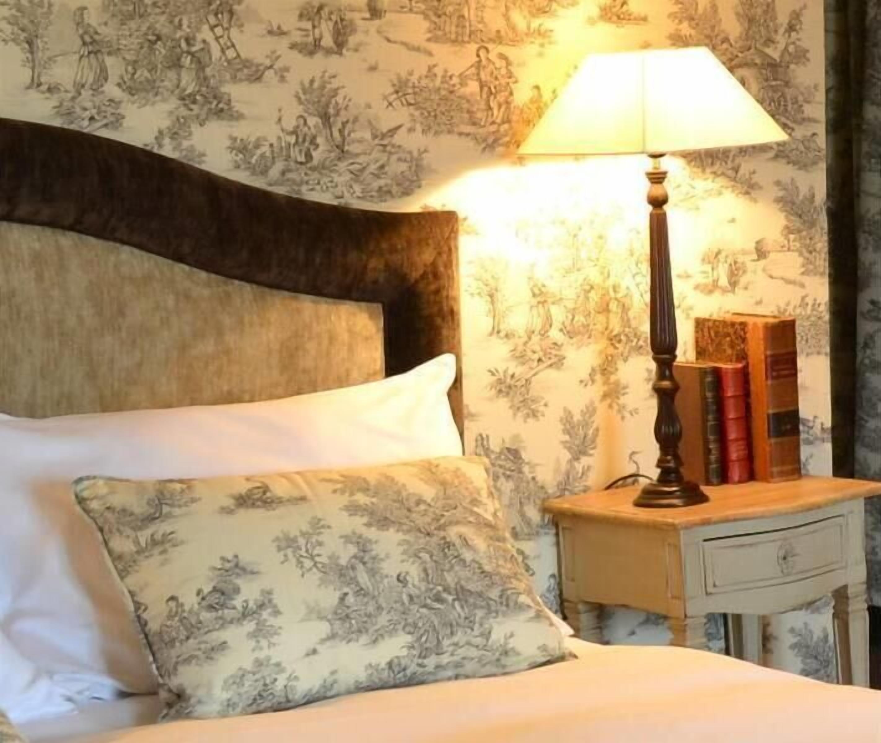 Een net opgemaakt bed met een bloemenkussen, een bedlampje en een nachtkastje met boeken en een lamp.
