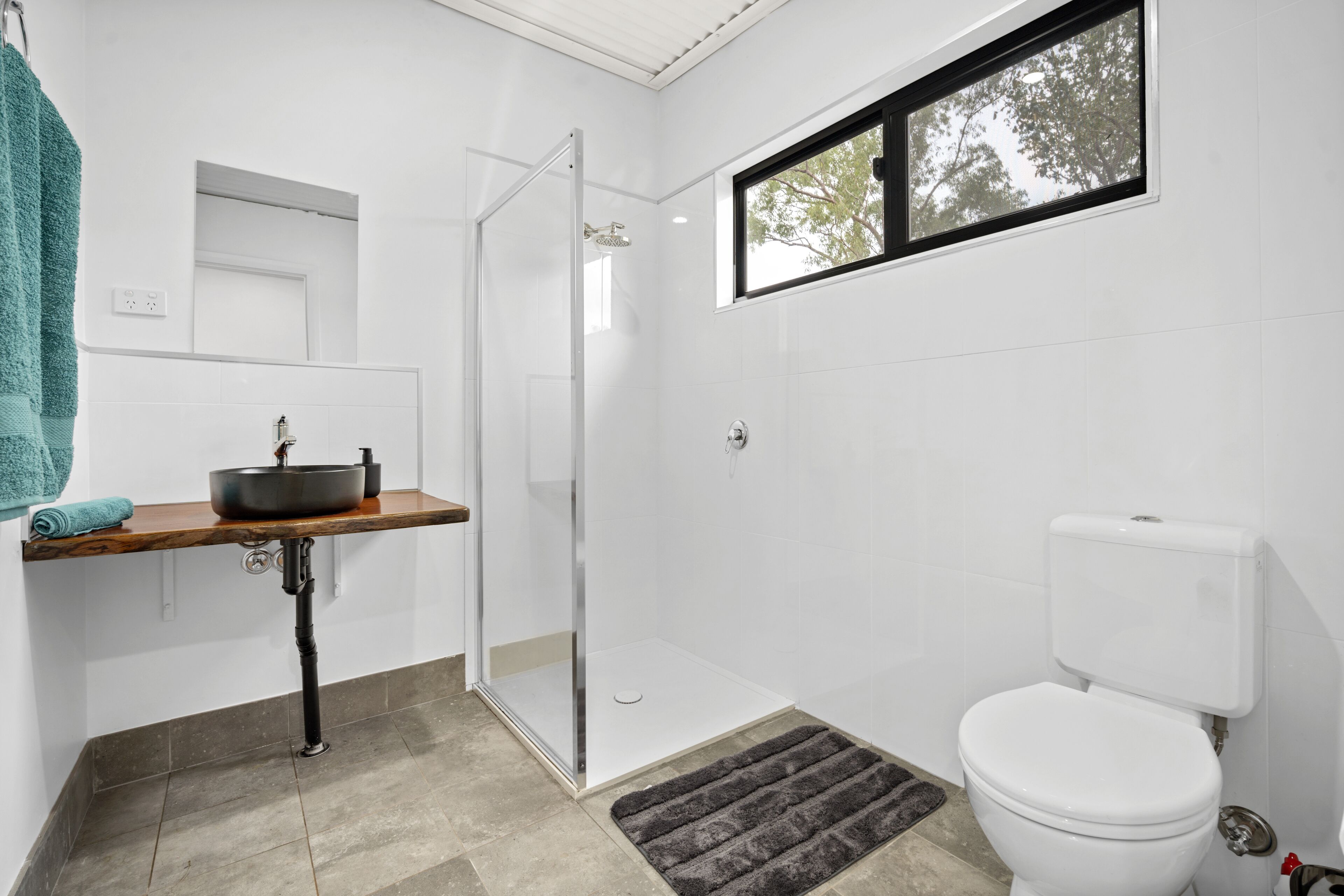 A bathroom with a glass shower enclosure, a toilet, a sink with a basin, and a window with a view of trees.