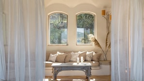 A cozy room with a sofa, a wooden coffee table, and two windows with views of greenery. - Villa Padulella 8 2 Emma Villas (Portoferraio)