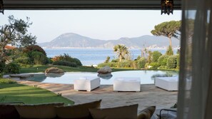 A poolside area with a view of the sea and mountains, featuring a small pool, white lounge chairs, and a well-maintained garden.