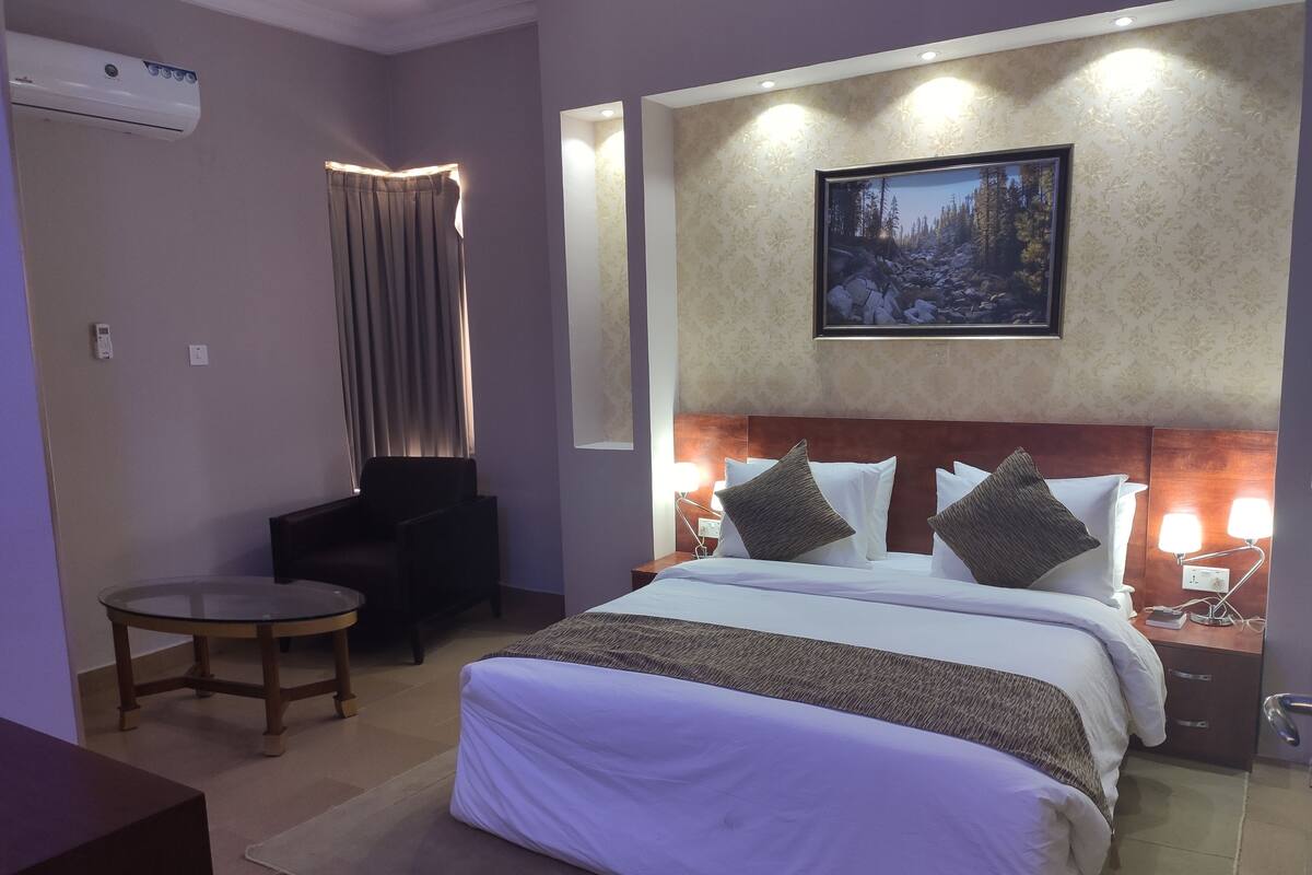 Habitación de hotel con una cama grande, una mesita redonda pequeña, una silla, una mesita de noche con lámparas y un cuadro en la pared.