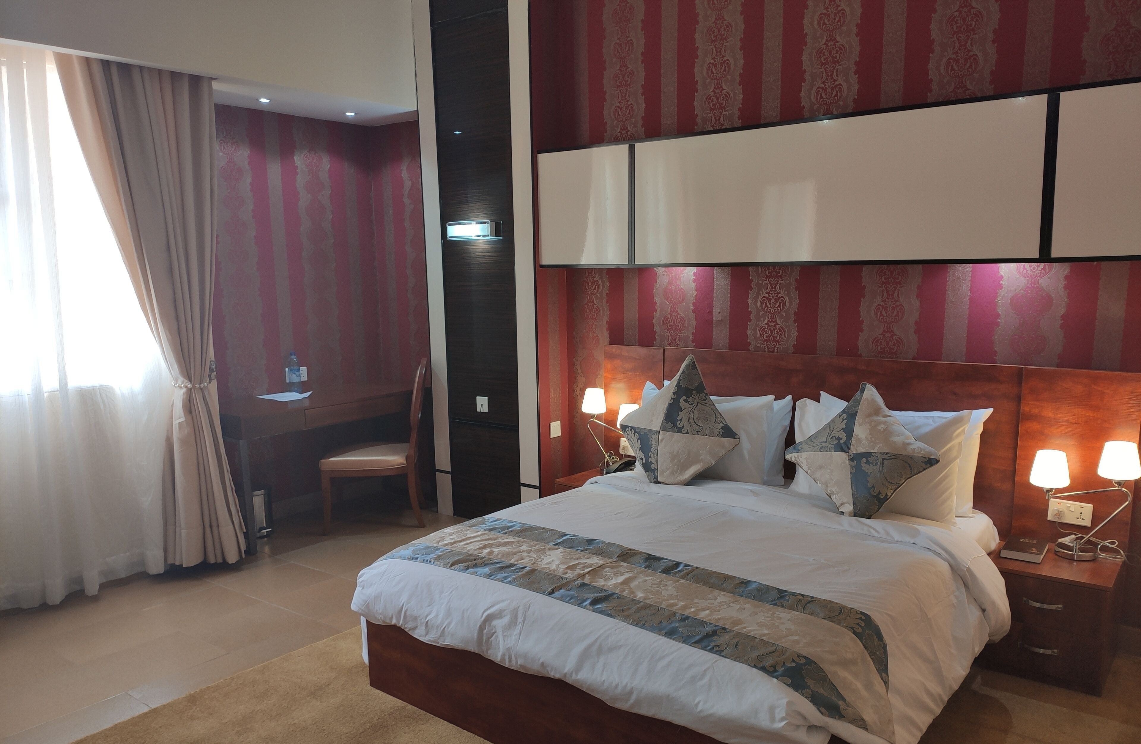A hotel room with a large bed, bedside lamps, a wooden headboard, and a desk with a chair.