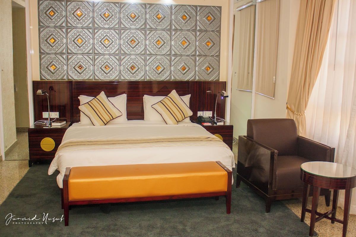 Habitación de hotel con una cama grande, un cabecero de madera, un sillón de cuero marrón, una mesita redonda pequeña y un panel decorativo en la pared.