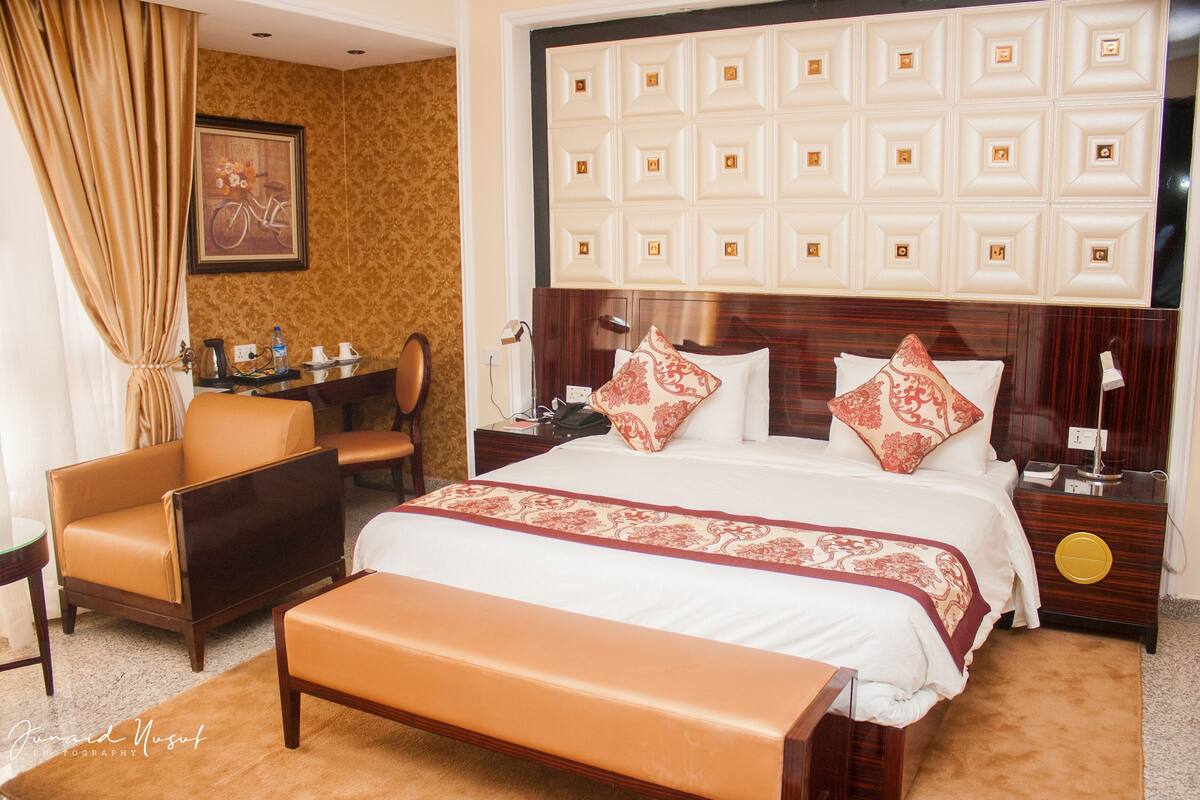 Habitación de hotel con una cama grande, un cabecero de madera, un escritorio con silla, un banco y un cuadro en la pared.