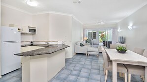 A modern kitchen with a central island, a dining area with a table and chairs, and a view of the outdoors through a glass door. - Alassio 1 Bedroom Ground Floor Pool Access 104/1 (Palm Cove)