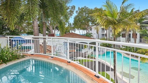 A swimming pool with a white railing, surrounded by palm trees and a building with balconies. - Alassio 1 Bedroom Ground Floor Pool Access 104/1 (Palm Cove)