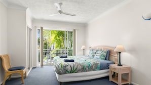 A bedroom with a bed, a chair, a nightstand, a ceiling fan, and a view of greenery through a sliding door. - 3 Bedroom Apartment 208 in Alassio Palm Cove (Cairns)