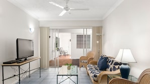 A living room with a television on a stand, a glass coffee table, wicker furniture, and a ceiling fan. - 3 Bedroom Apartment 208 in Alassio Palm Cove (Cairns)