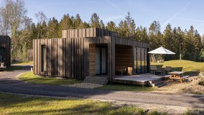Una cabaña moderna de madera con un balcón, mobiliario exterior y una zona cubierta con sombrilla.