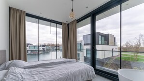 A modern bedroom with a large bed, a glass-enclosed balcony, and a view of a modern building across a body of water. - Waterfront holiday home in Arnemuiden (Arnemuiden)