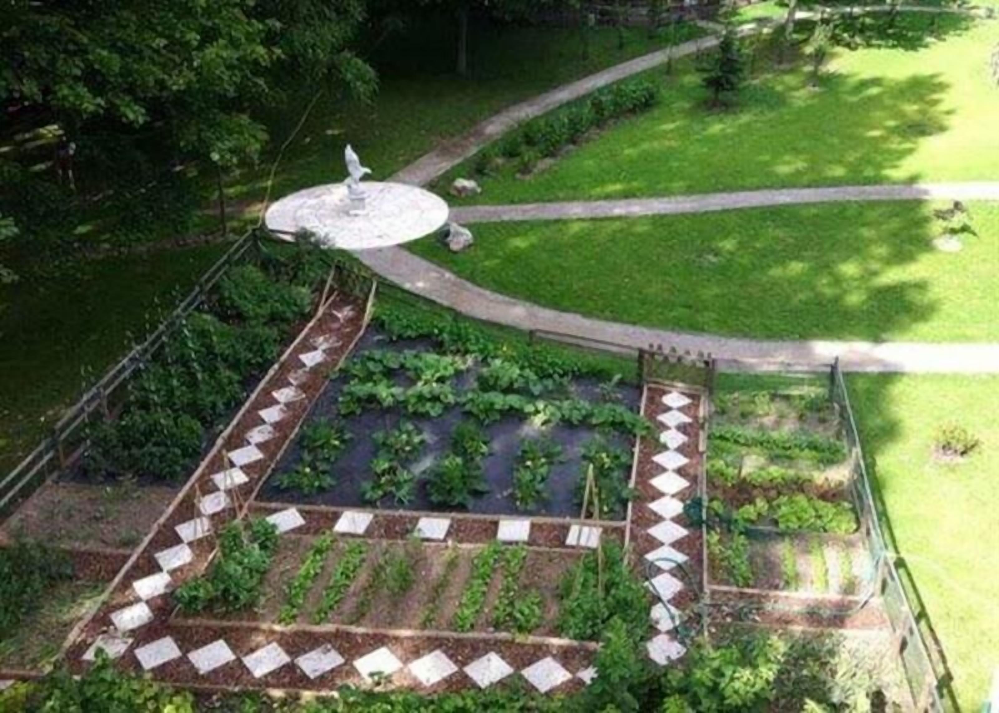 A garden with a central fountain, a paved walkway, and raised garden beds with plants.