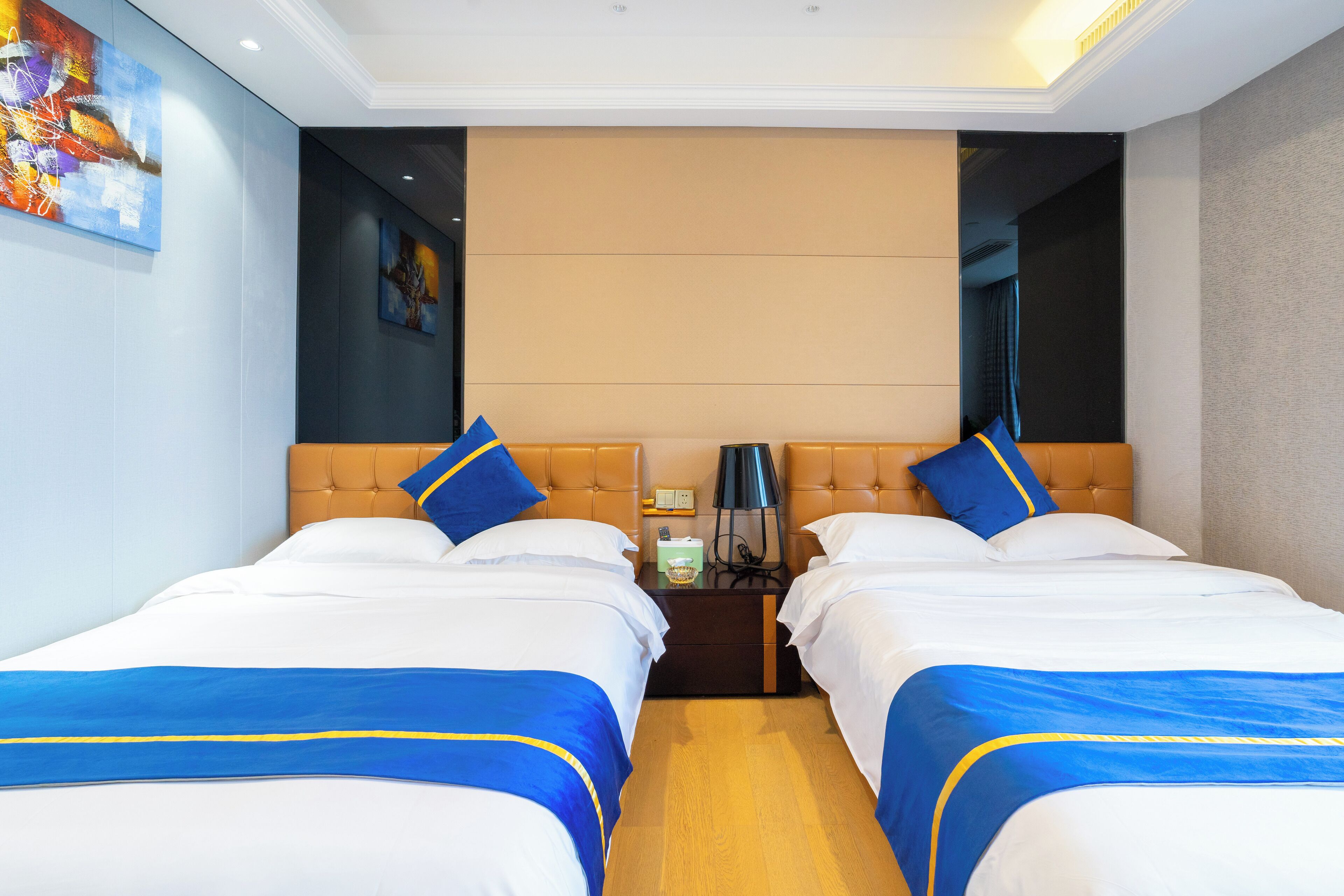 A hotel room with two beds, each with a blue and white bedspread and matching pillows. There is a nightstand between the beds with a lamp and a small table. The room has a modern design with a neutral color palette and artwork on the walls.
