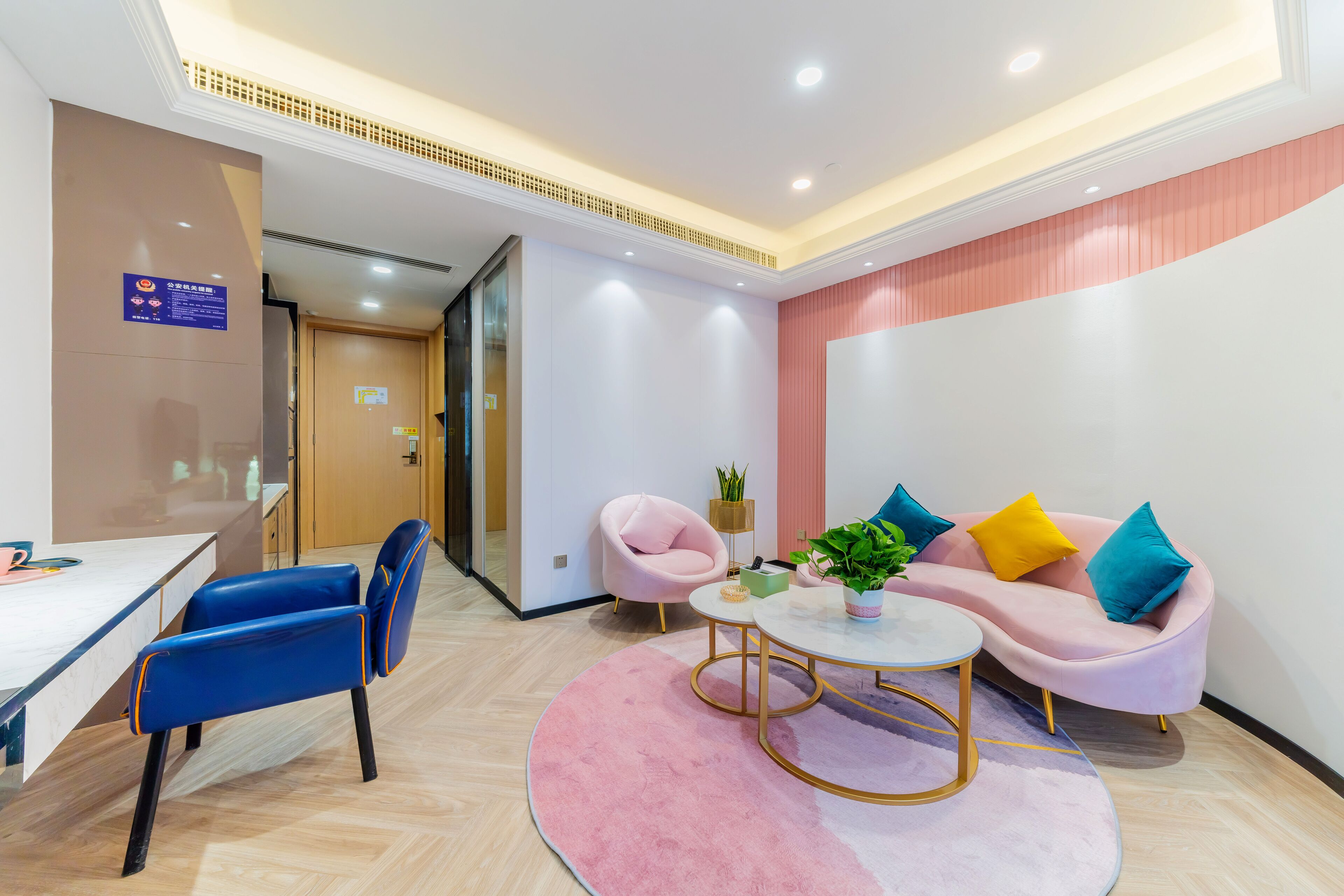 Un hall d’hôtel moderne avec un comptoir d’accueil, un canapé rose agrémenté de coussins colorés, un fauteuil bleu et une petite table ronde sur laquelle est posée une plante.