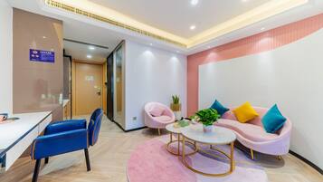 A modern hotel lobby with a reception desk, a pink sofa with colorful cushions, a blue chair, and a small round table with a plant.