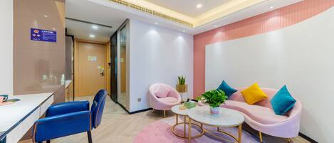 Un moderno vestíbulo de hotel con una recepción, un sofá rosa con cojines coloridos, una silla azul y una pequeña mesa redonda con una planta.
