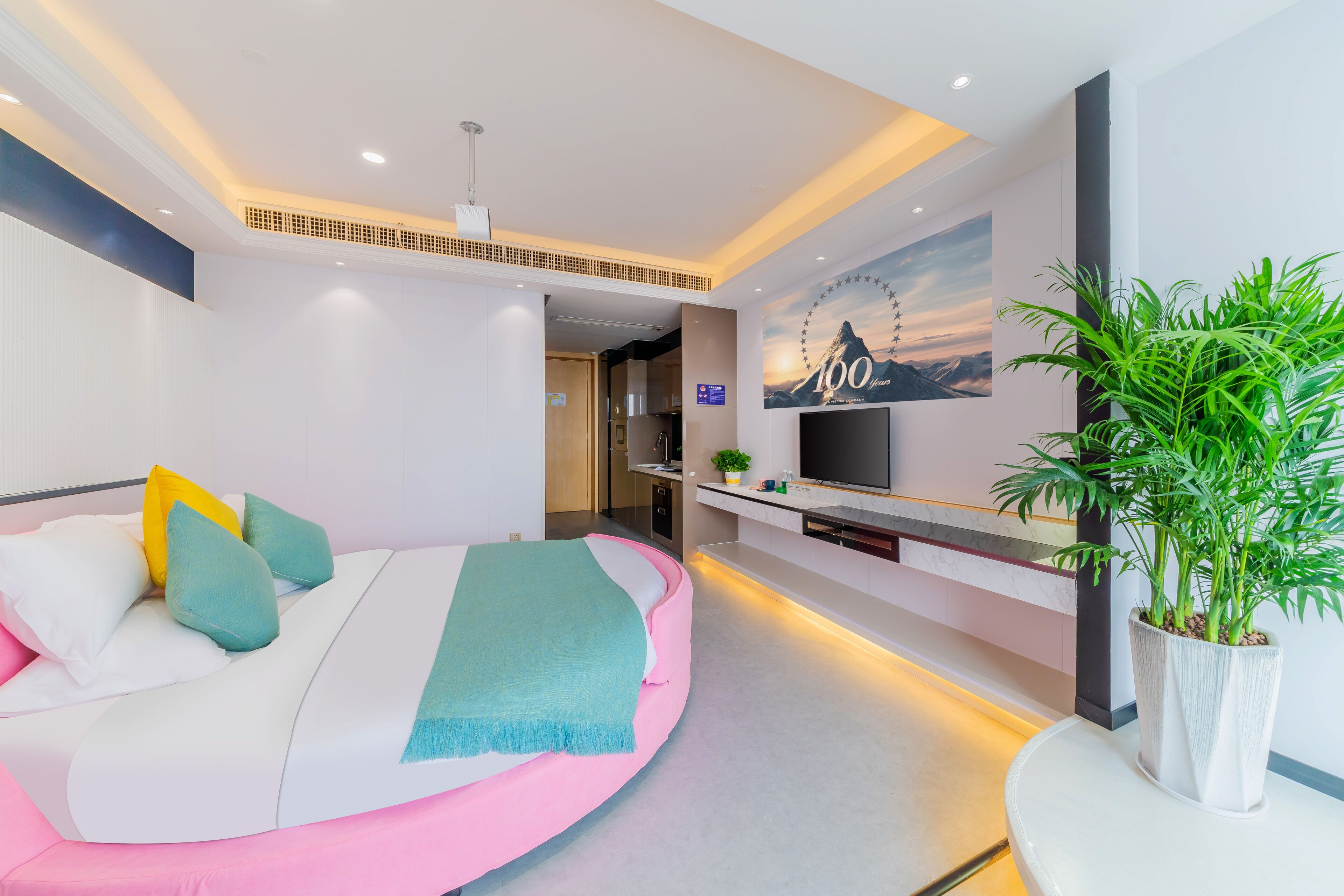 A modern hotel room with a curved bed, a television, a plant, and a mural on the wall.