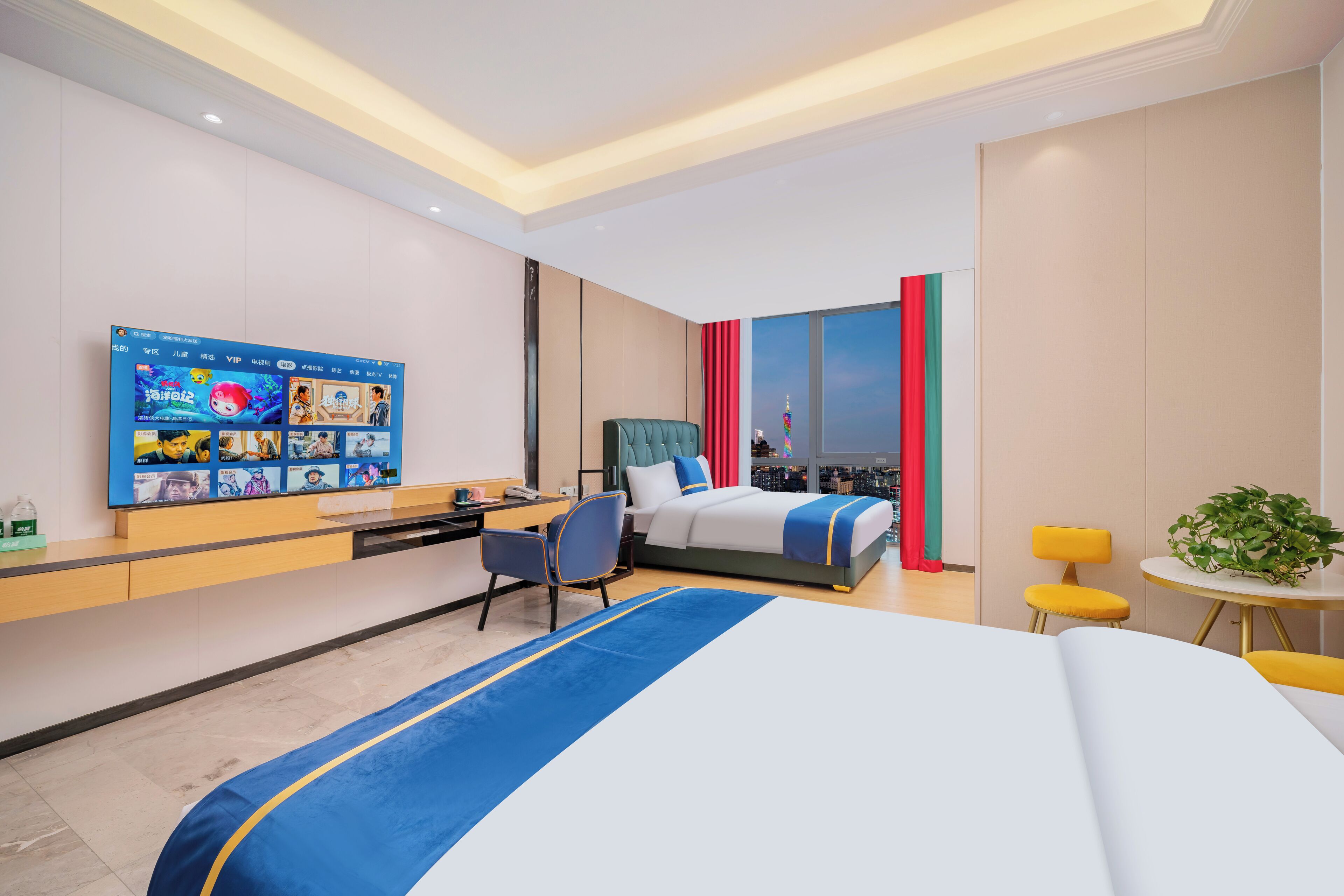 A modern hotel room with a large bed, a desk with a chair, a small table with a plant, and a TV displaying various channels.