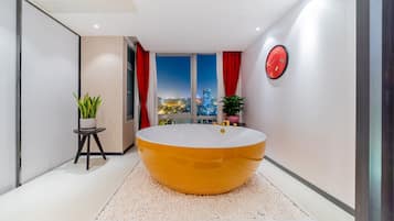 A modern bathroom with a large oval bathtub, a potted plant, and a city view through a large window.