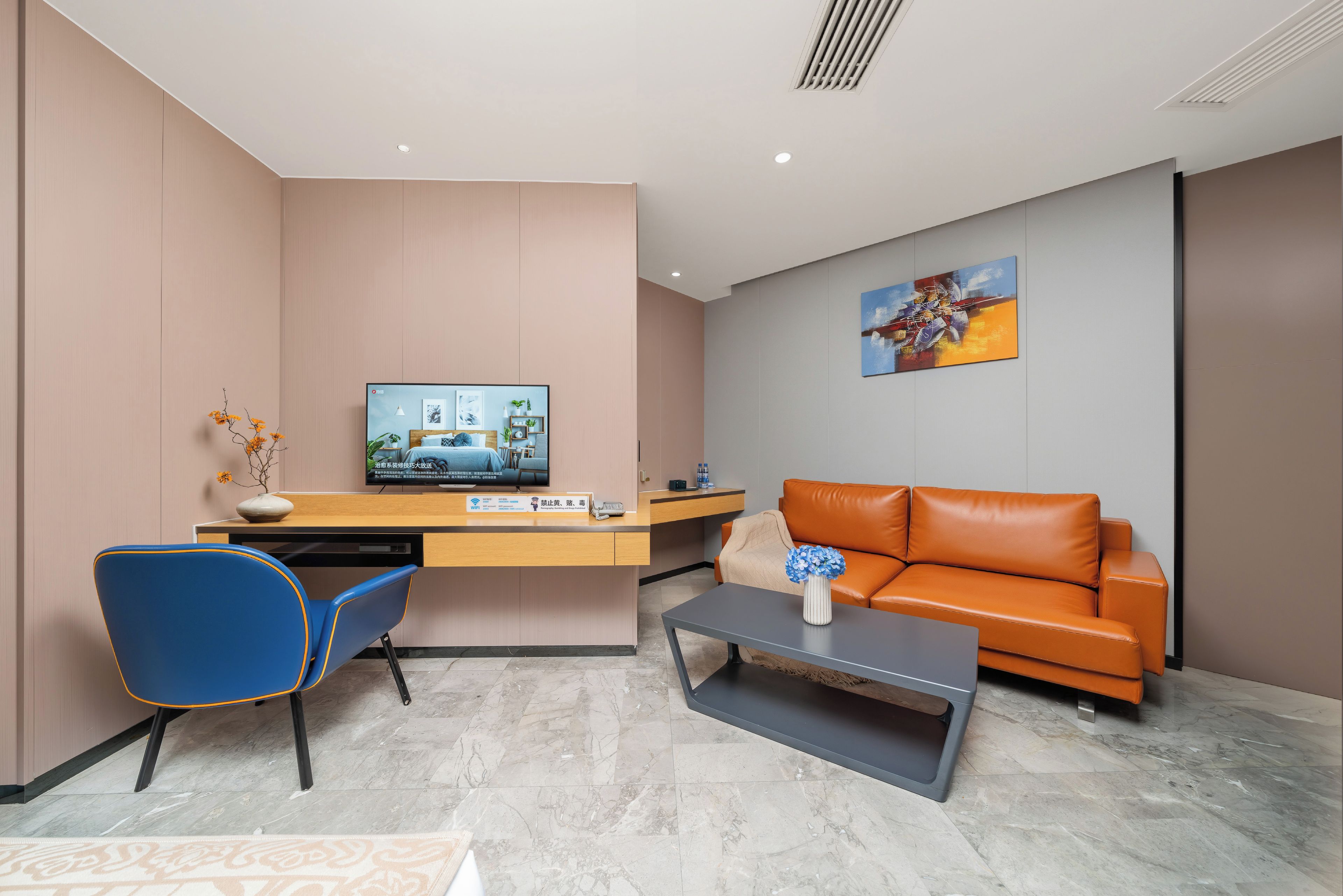 Una habitación de hotel moderna con un escritorio, una silla azul, un sofá naranja, una mesa de centro y un televisor.