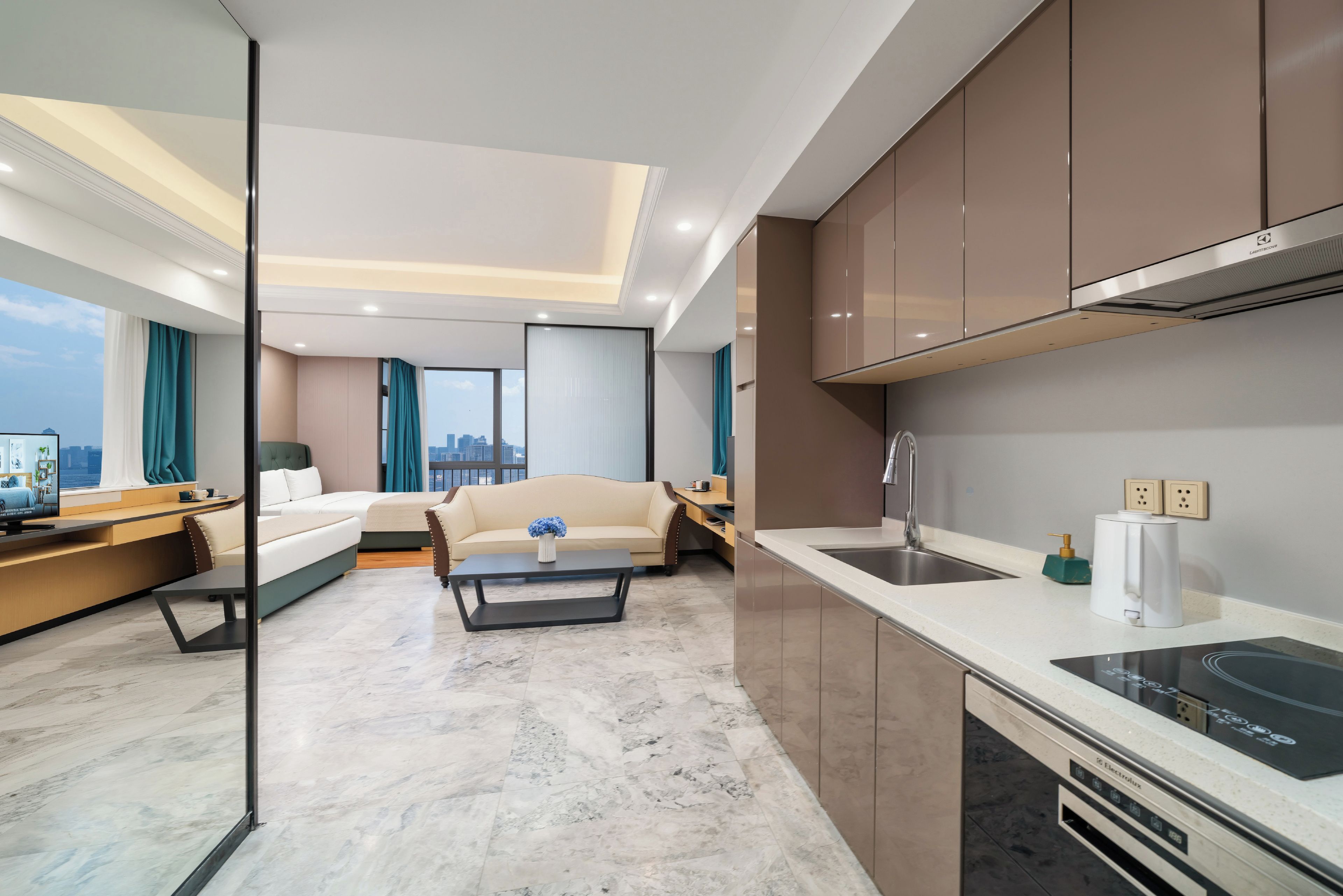 A modern hotel room with a sofa, a coffee table, a TV, a kitchen area with a sink and stove, and a city view.