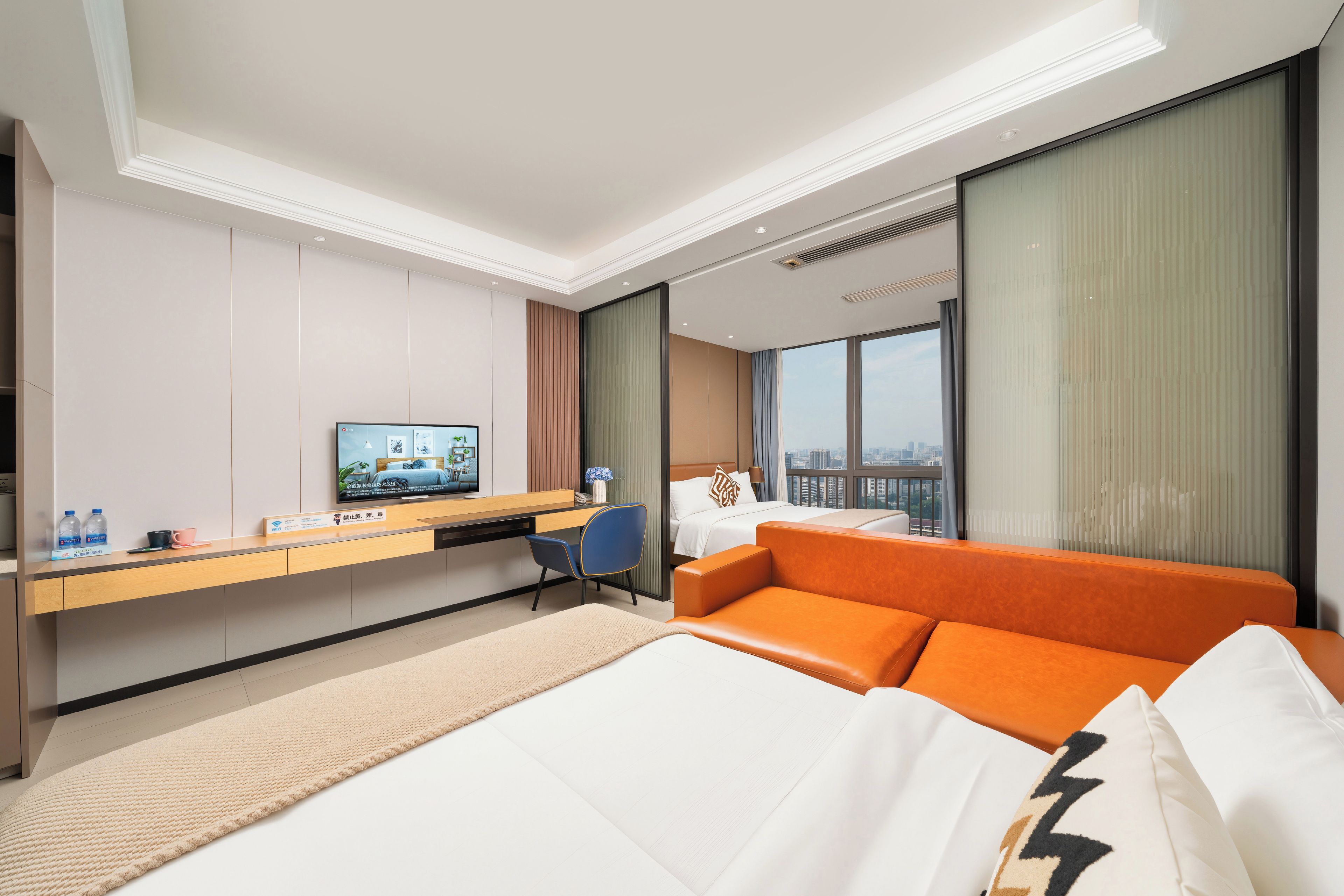 Habitación de hotel con una cama, un escritorio con computadora, una silla, un televisor y un amplio ventanal con vista a la ciudad.