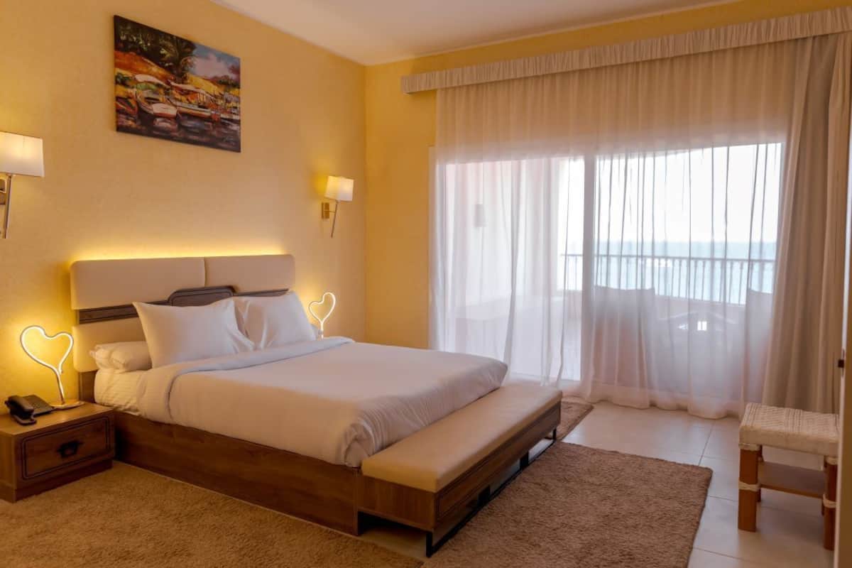 Une chambre d’hôtel avec un grand lit, des lampes de chevet, un banc, un tableau au mur et une vue à travers des rideaux légers.