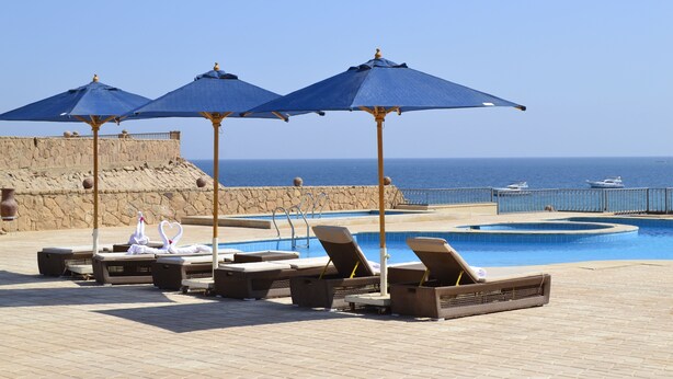 Un espace aménagé au bord de la piscine, avec des chaises longues et des parasols, un mur en pierre et une vue sur la mer.