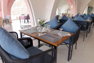 Un comedor con mesas dispuestas para dos, con sillas acolchadas de color azul y un florero central con flores.