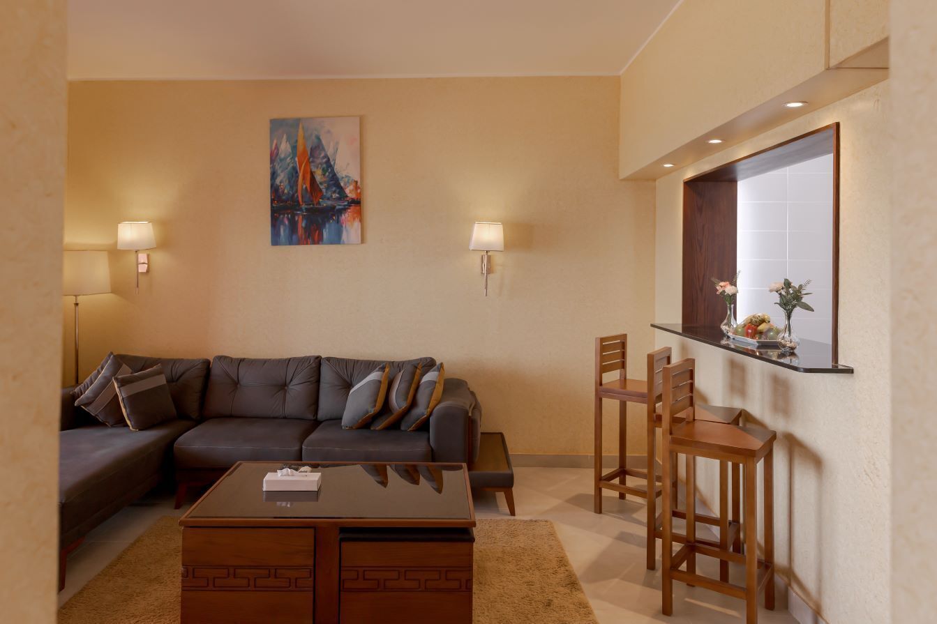 A living room with a sofa, coffee table, and chairs. There is a painting on the wall and a window with a view.