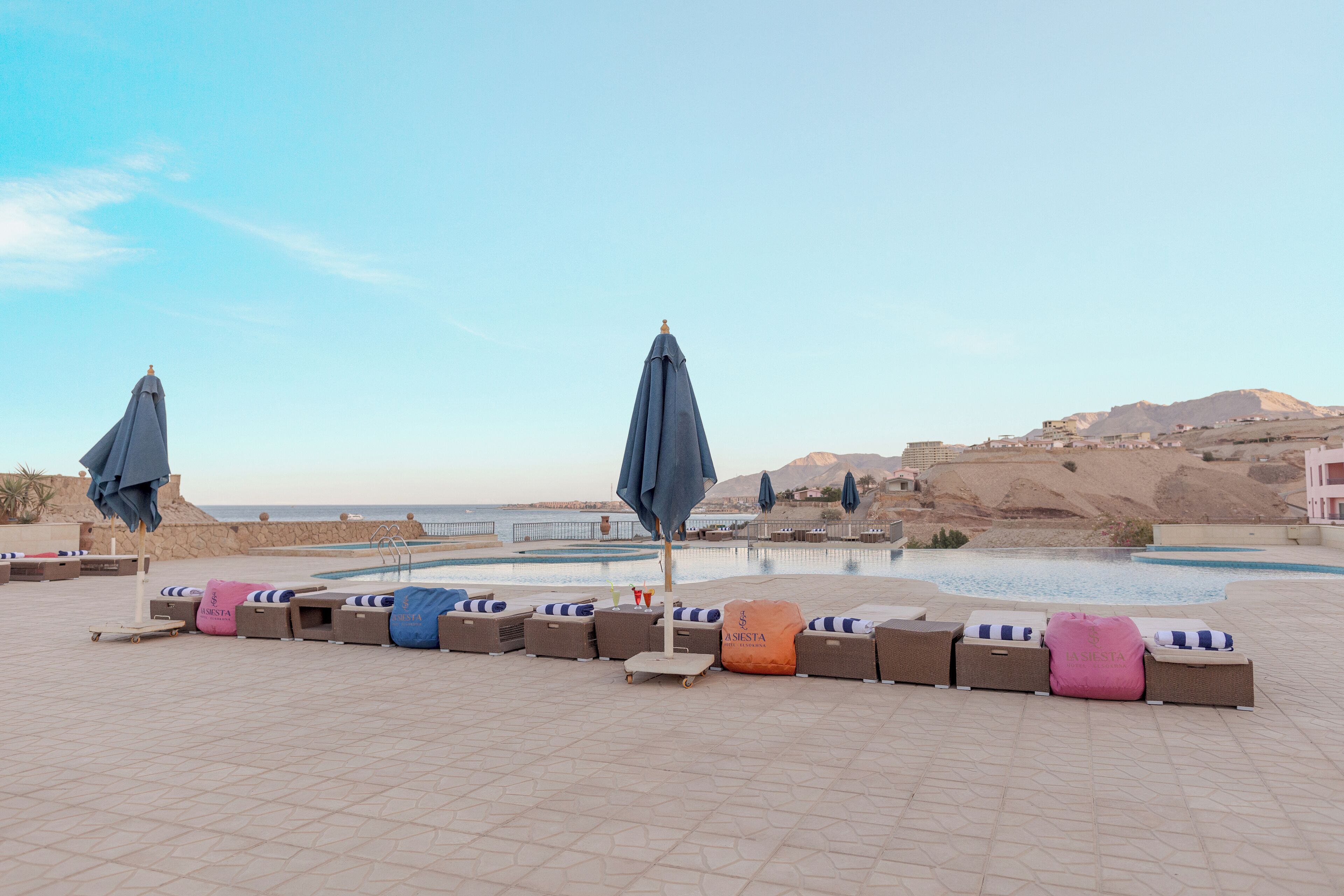 Zona junto a la piscina con sillones de descanso y sombrillas, cielo despejado y una vista distante de un paisaje montañoso.