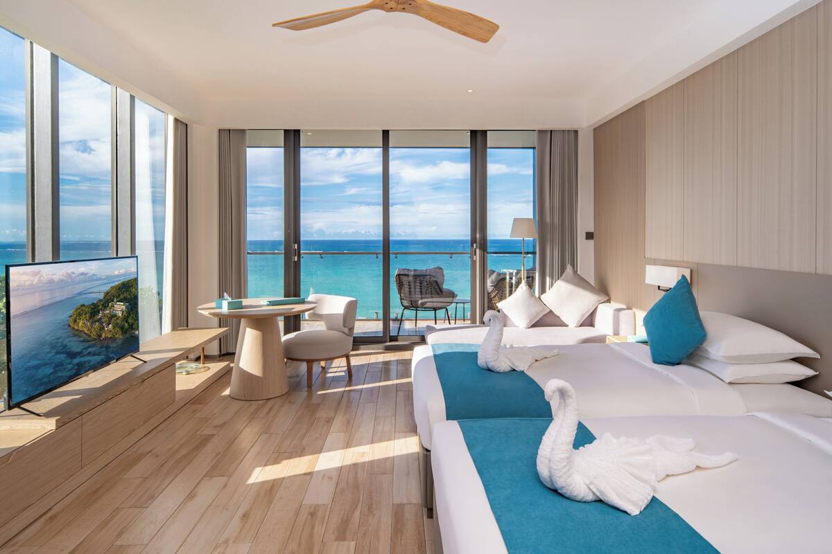 Ein modernes Hotelzimmer mit einem großen Bett, einem Fernseher, einem Balkon mit Meerblick und einem Essbereich.