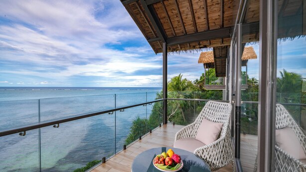 Een balkon met glazen hekwerk, uitzicht op zee, rieten stoelen, een ronde tafel met fruit en een rieten dakconstructie.