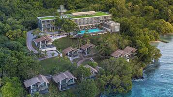 Vista aérea de un resort con piscina, varias construcciones y abundante vegetación rodeando un mar azul cristalino.