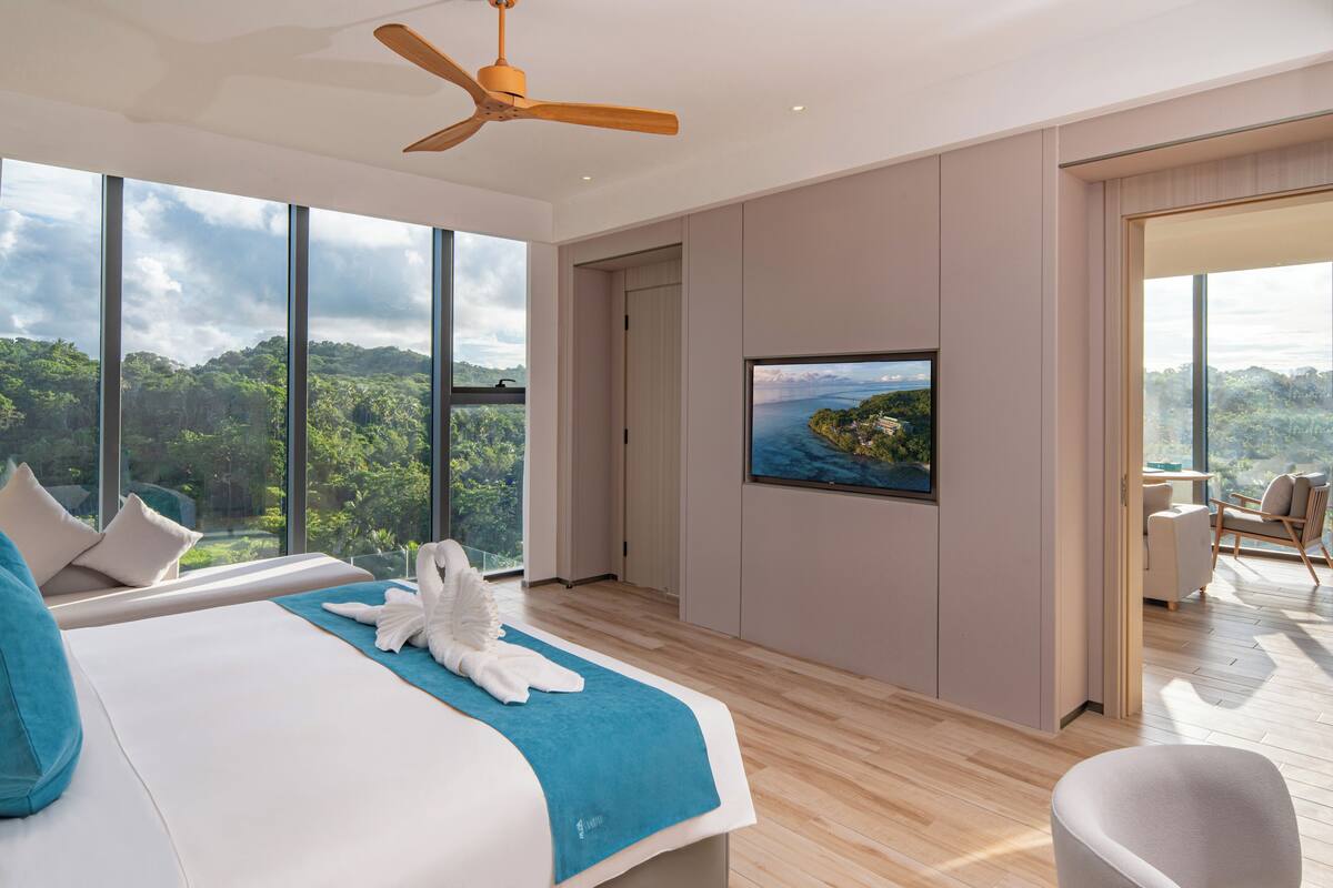 Ein modernes Hotelzimmer mit einem großen Bett, einem Deckenventilator, einem Fernseher und Blick auf eine gebirgige Landschaft.