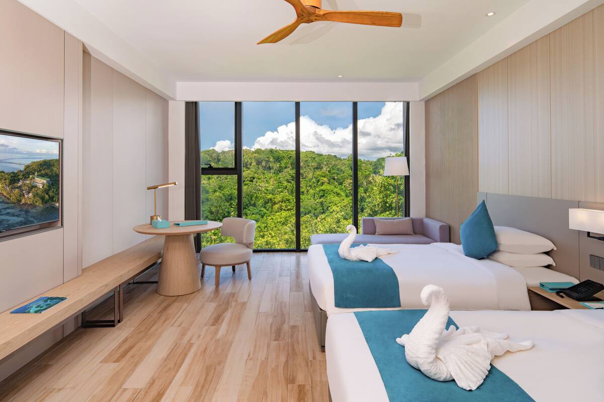 Ein modernes Hotelzimmer mit großem Fenster, einem Bett mit blauer Decke, einer Bank, einem Schreibtisch und einem Deckenventilator.