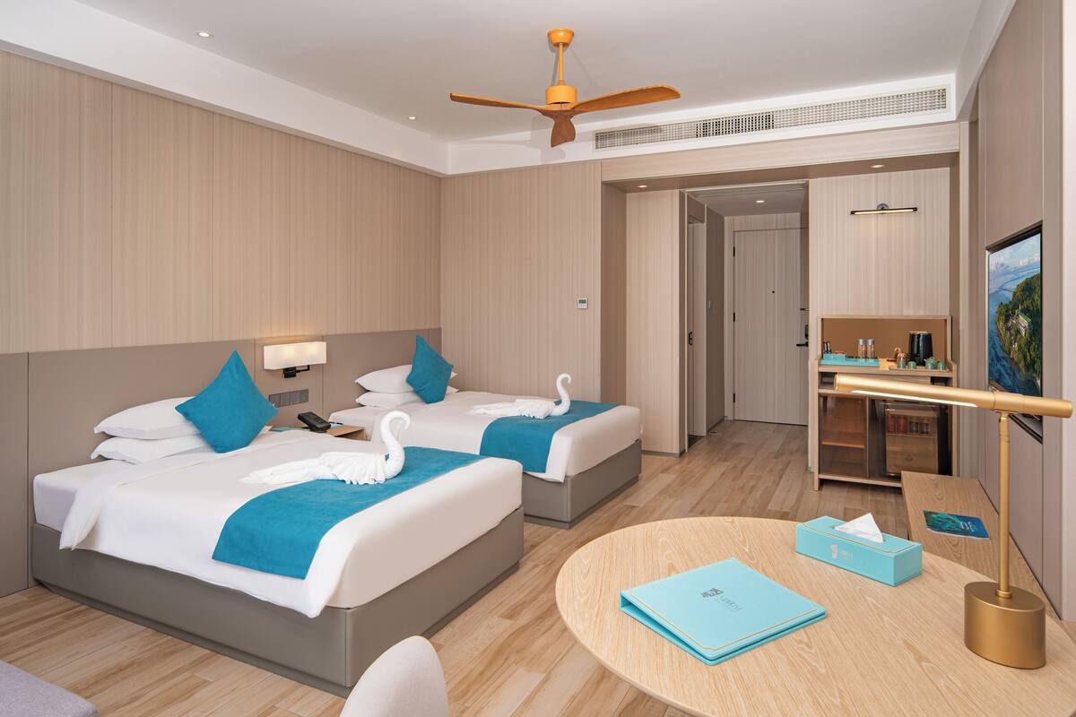 Ein modernes Hotelzimmer mit zwei Betten, einem Schreibtisch, einem runden Tisch und einem Deckenventilator.
