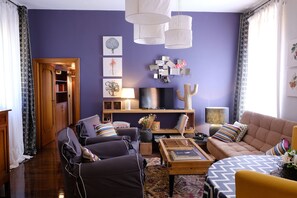 Ein Wohnzimmer mit einer Couch, Sesseln, einem Couchtisch und einem Fernseher. Außerdem befindet sich dort ein Bücherregal und ein dekoratives Wandbild.